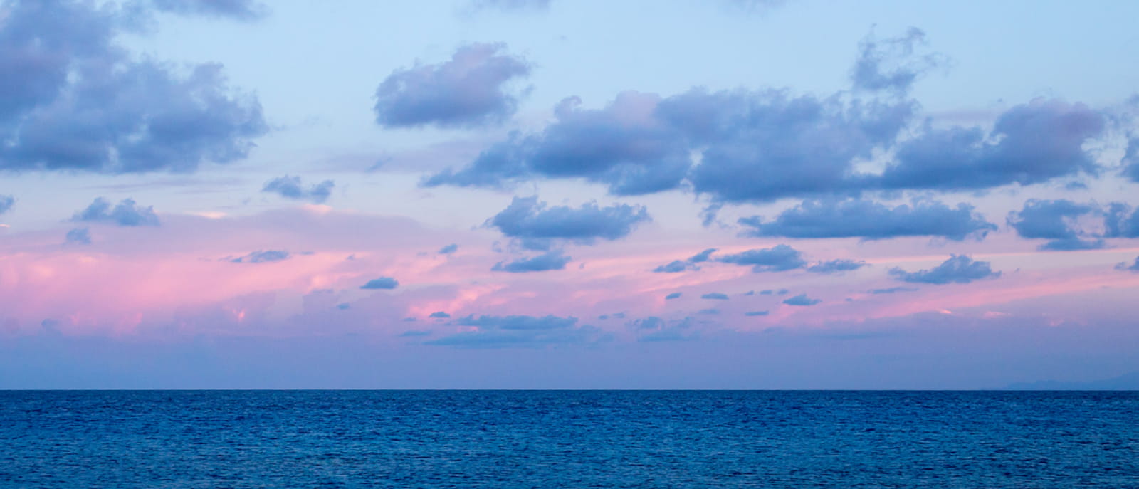 Hero image of ocean at sunset with a sky displaying shades of pink and blue, scattered with clouds.