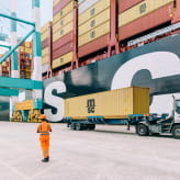 An MSC cargo ship being loaded with containers at a port, with a worker and trucks handling MSC-branded containers under large cranes.