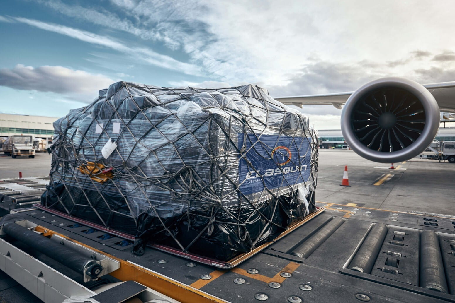 Cargo wrapped in plastic and secured with netting is loaded onto an airport conveyor belt, with an airplane engine and terminal in the background.