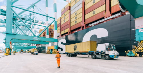 An MSC cargo ship being loaded with containers at a port, with a worker and trucks handling MSC-branded containers under large cranes.