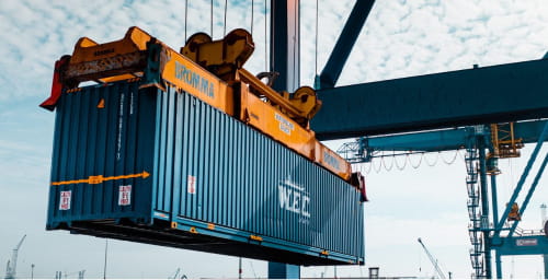 A WEC Lines shipping container being lifted by a crane at a port, with industrial equipment and a clear sky in the background.