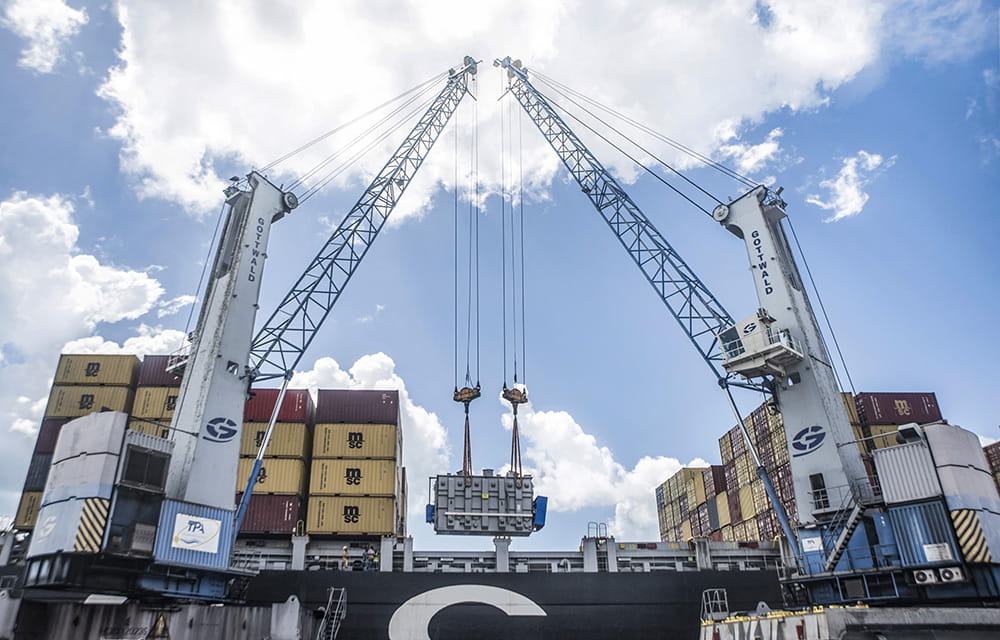 A large crane hoists a transformer onto a cargo ship at a bustling port